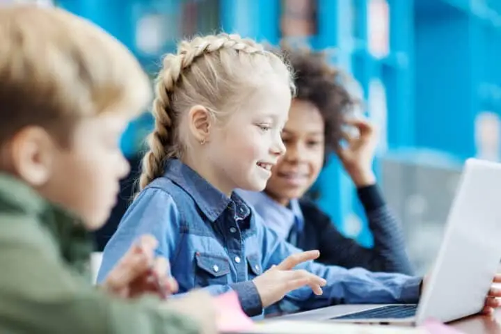 Group of three elementary students sitting at desk in school library and browsing Internet on laptop